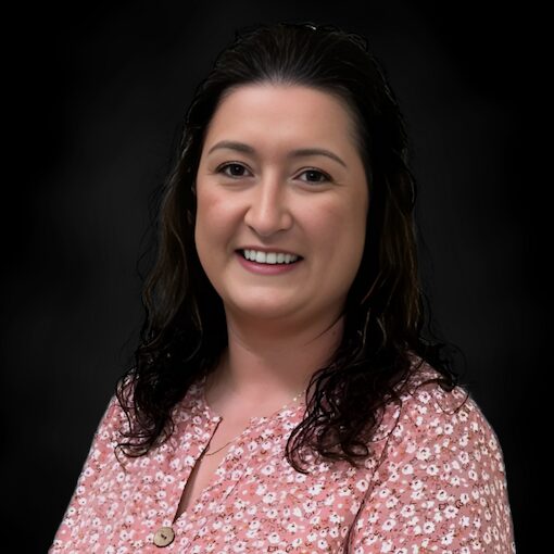 A woman with wavy dark hair smiles at the camera. She is wearing a pink, floral-patterned blouse and is posed in front of a plain, dark background.