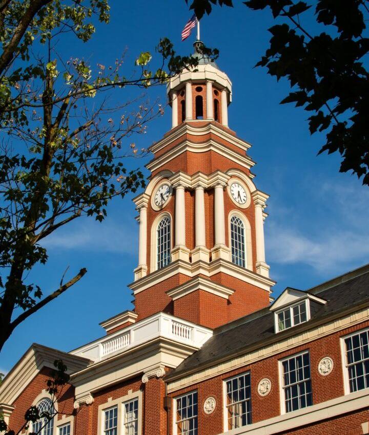 A tall red-brick clock tower rises against a clear blue sky, framed by leafy tree branches. The building has white trim, arched windows, and a small American flag atop the tower.