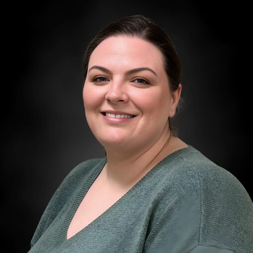 A woman with fair skin and dark hair, pulled back, is smiling at the camera. She is wearing a green textured top and is set against a dark, softly blurred background.