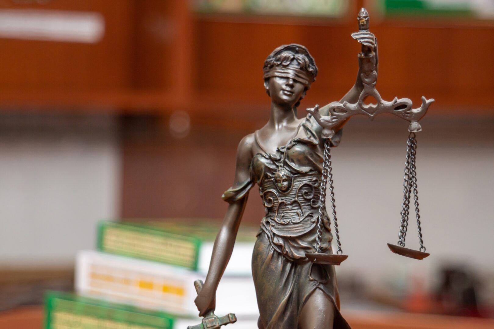 A close-up of a bronze Lady Justice statue holding balanced scales and a sword, with a blindfold over her eyes. Blurred books and shelves appear in the background, symbolizing The Push for Guardian ad Litem Reform in West Virginia Family Courts.