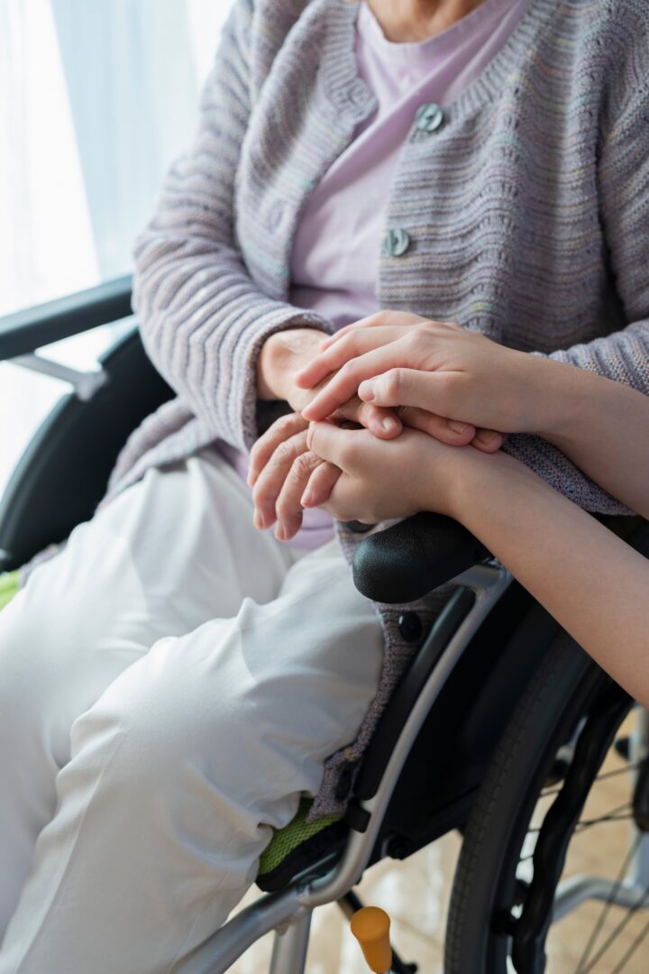 An older adult in a wheelchair is gently holding hands with another person, conveying support and comfort. The focus is on their hands and laps, with soft natural light in the background.