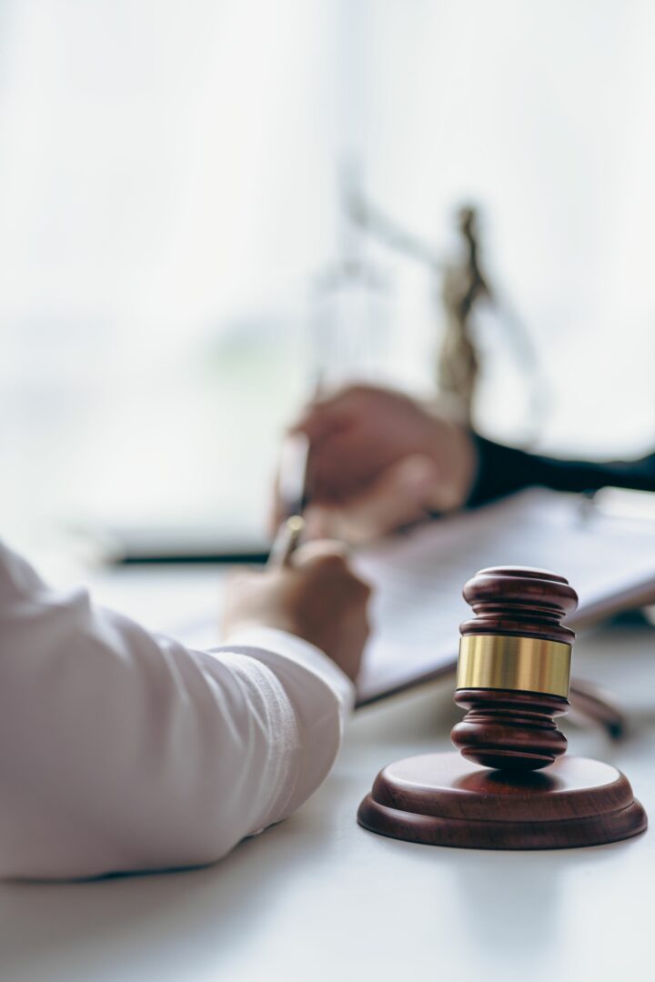 Close-up of a wooden judge’s gavel on a desk, with two people in the background discussing and signing documents, blurred out, suggesting a legal or courtroom setting. Close-up of a wooden judge’s gavel on a desk, with two people in the background discussing and signing documents, blurred out, suggesting a legal or courtroom setting.