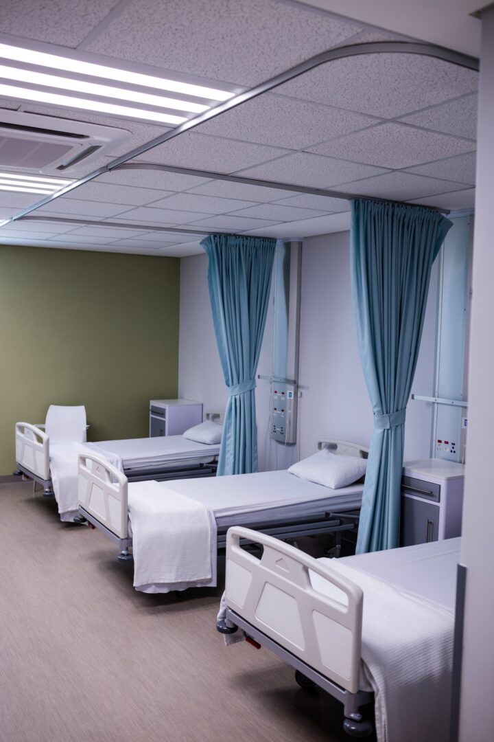 Three empty hospital beds with white sheets and pillows are separated by blue privacy curtains in a clean, well-lit hospital room. A white chair and medical equipment are visible near the beds. Three empty hospital beds with white sheets and pillows are separated by blue privacy curtains in a clean, well-lit hospital room. A white chair and medical equipment are visible near the beds.