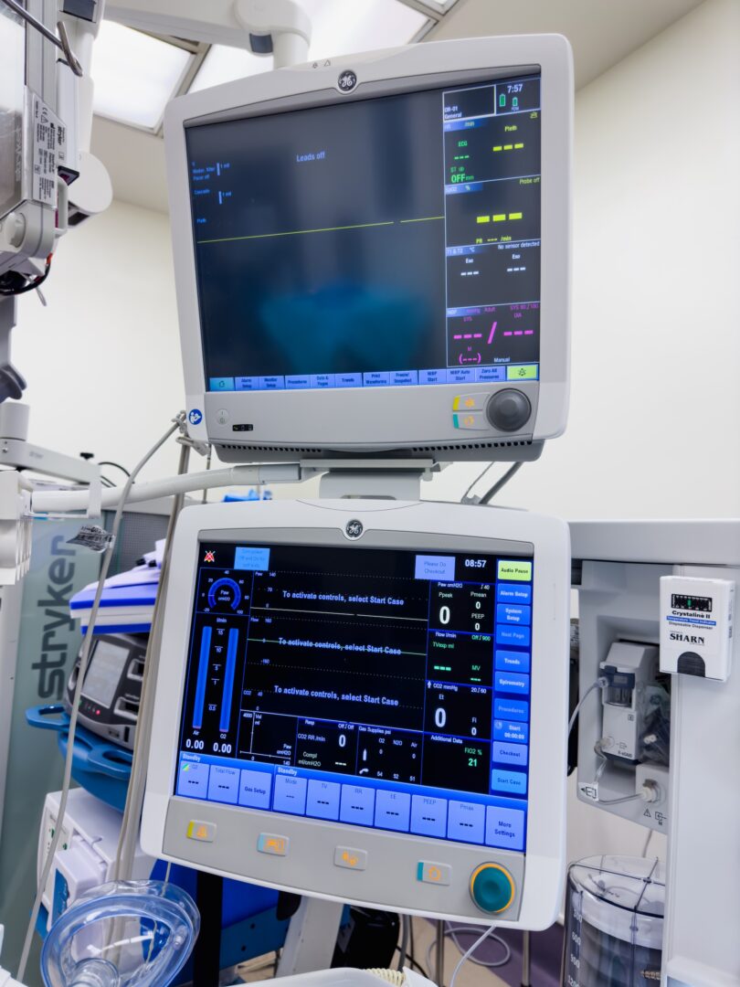 Two medical monitors in a hospital setting display patient vital signs and respiratory data. The screens show various colorful graphs, numbers, and status information, surrounded by medical equipment and cables. Two medical monitors in a hospital setting display patient vital signs and respiratory data. The screens show various colorful graphs, numbers, and status information, surrounded by medical equipment and cables.