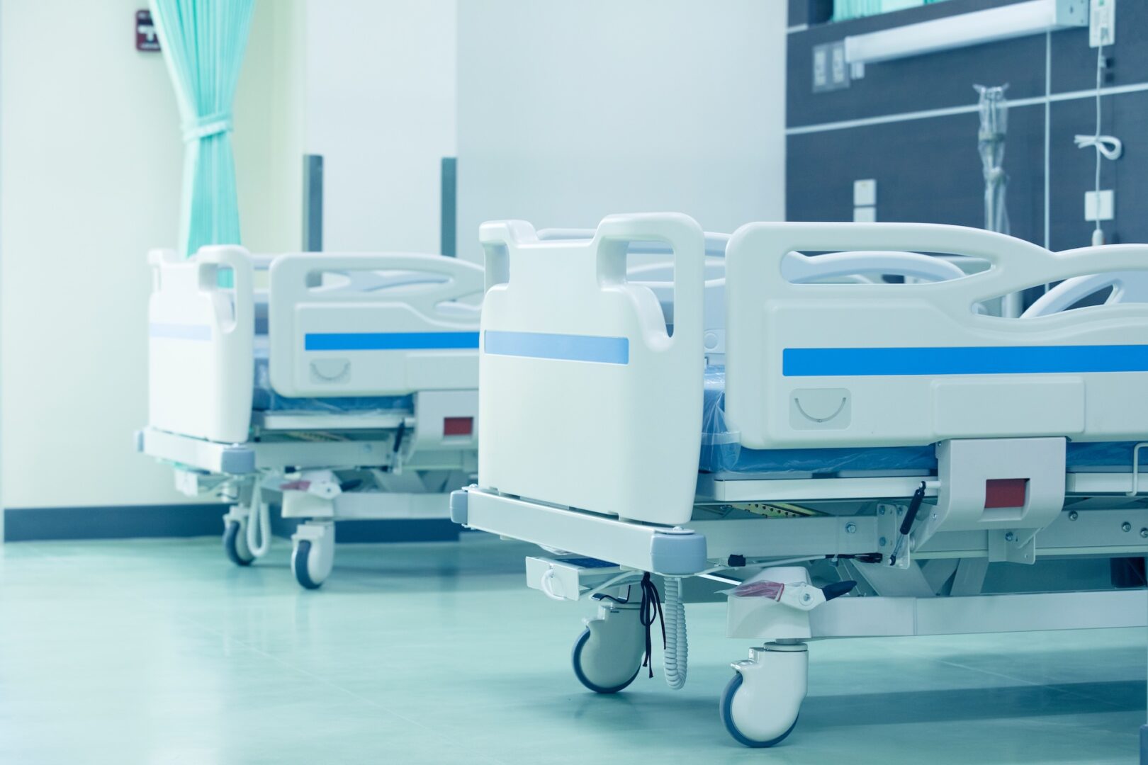Two empty hospital beds with blue accents are positioned in a clean, bright hospital room. Curtains and medical equipment are visible in the background.