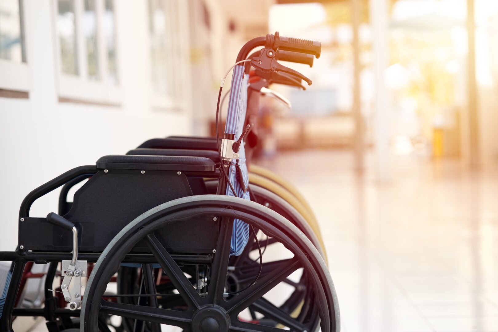 A row of empty wheelchairs is lined up in a bright, sunlit hallway with a reflective floor, next to a wall with windows.
