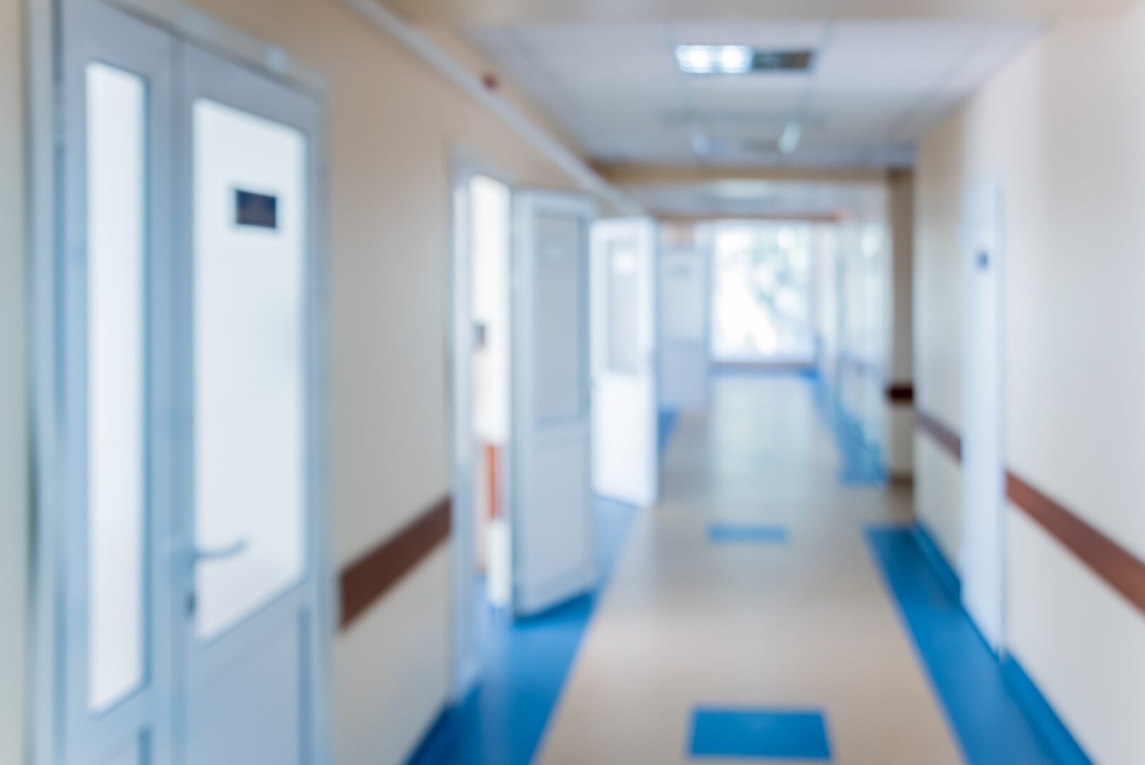 A blurred image of a hospital or clinic hallway with cream-colored walls, blue flooring, and several white doors, some of which are open. Fluorescent lights are visible on the ceiling.