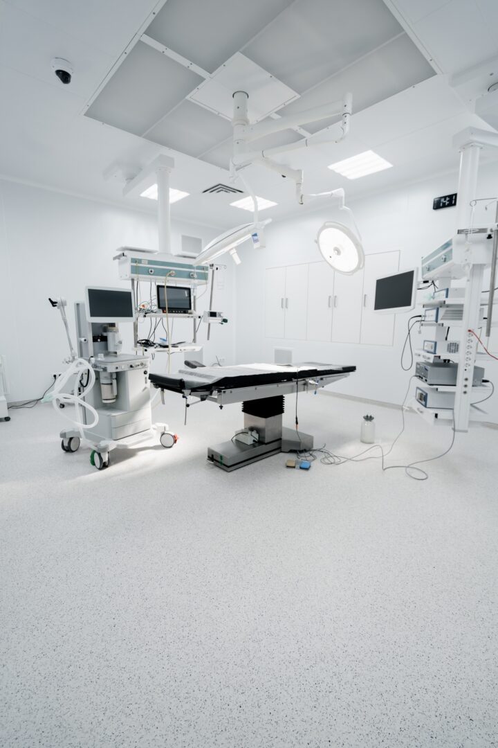 A modern, empty operating room with a surgical table in the center, surrounded by medical equipment, monitors, overhead lights, and white walls and floors, creating a sterile environment. A modern, empty operating room with a surgical table in the center, surrounded by medical equipment, monitors, overhead lights, and white walls and floors, creating a sterile environment.