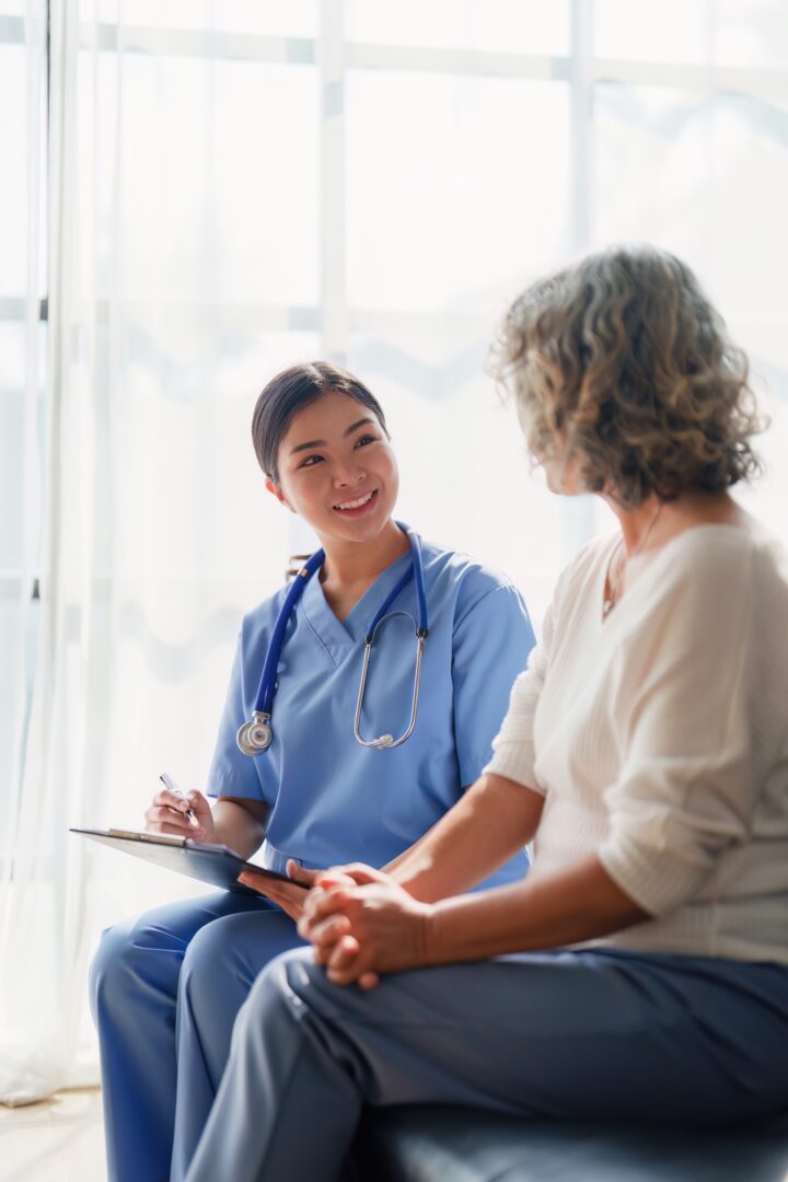 A smiling healthcare professional in blue scrubs with a stethoscope talks to an older woman in casual clothes, holding a clipboard in a bright, sunlit room.