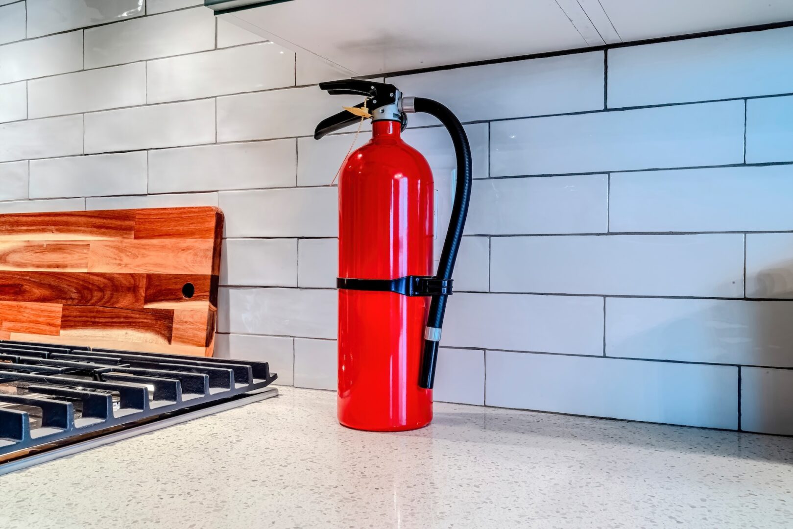 A red fire extinguisher is placed on a white kitchen countertop next to a stove with a wooden cutting board against a white tiled backsplash.