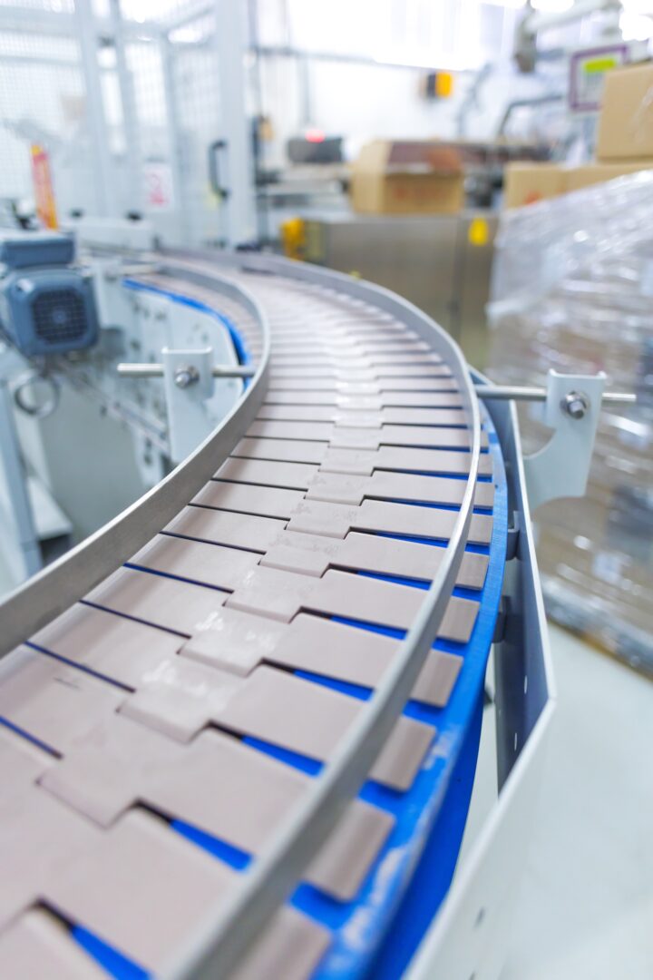A close-up of an empty, curved conveyor belt in a brightly lit industrial setting, with machinery and stacked boxes visible in the background. A close-up of an empty, curved conveyor belt in a brightly lit industrial setting, with machinery and stacked boxes visible in the background.