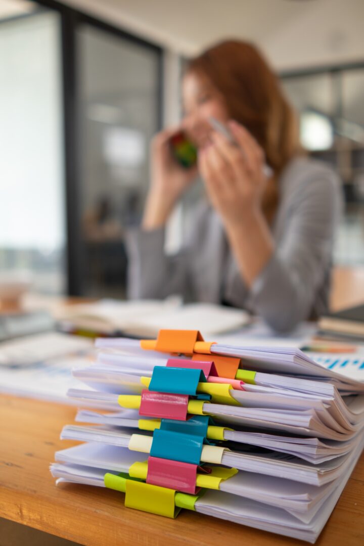 A stack of documents organized with colorful binder clips sits on a desk, while a person in the background is on the phone, appearing out of focus. A stack of documents organized with colorful binder clips sits on a desk, while a person in the background is on the phone, appearing out of focus.
