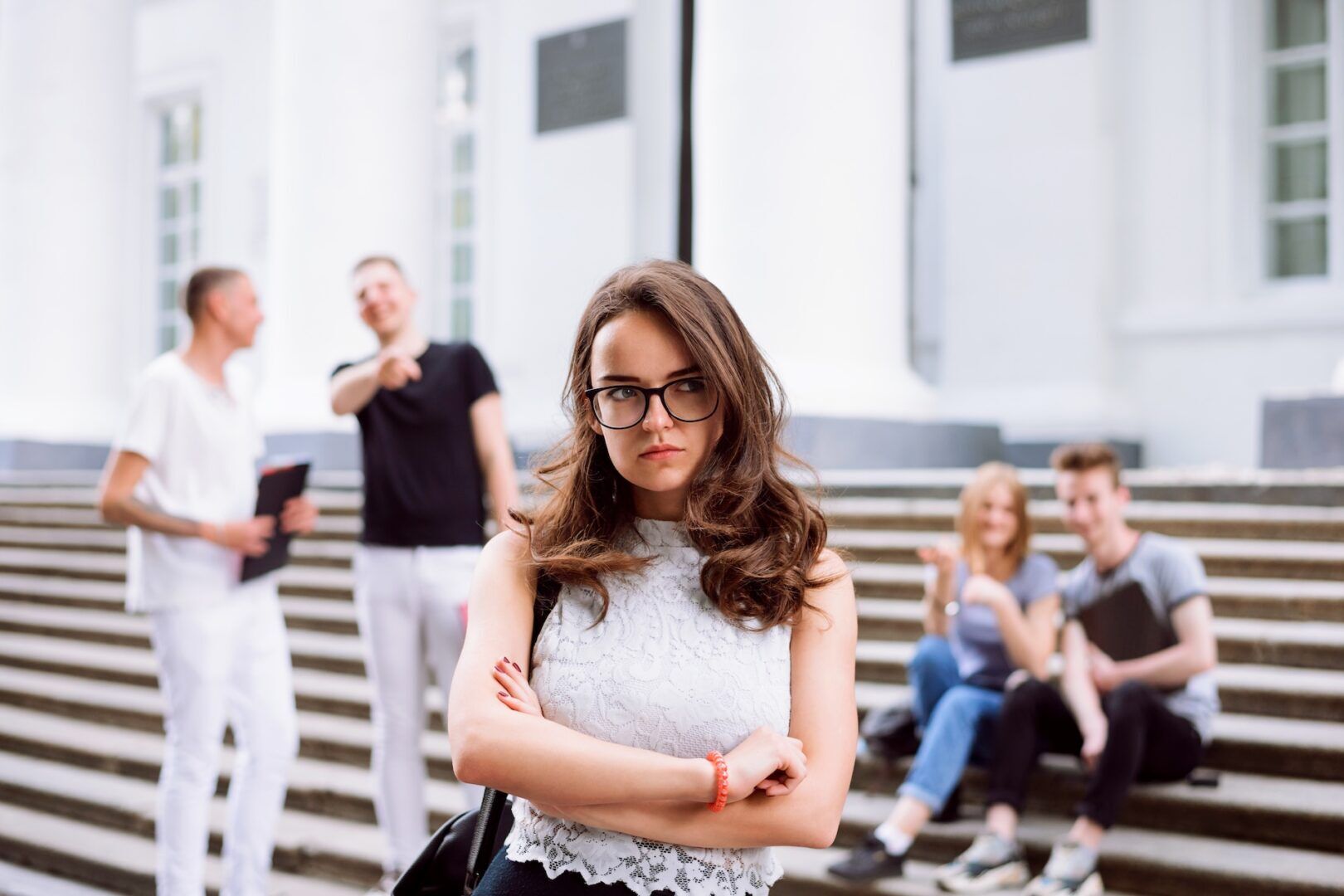 A young woman with glasses stands in front of a building, arms crossed and looking upset—capturing the tension seen in "How to Recognize and Respond to Workplace Discrimination in West Virginia." Behind her, two people laugh and point while others talk on the steps.