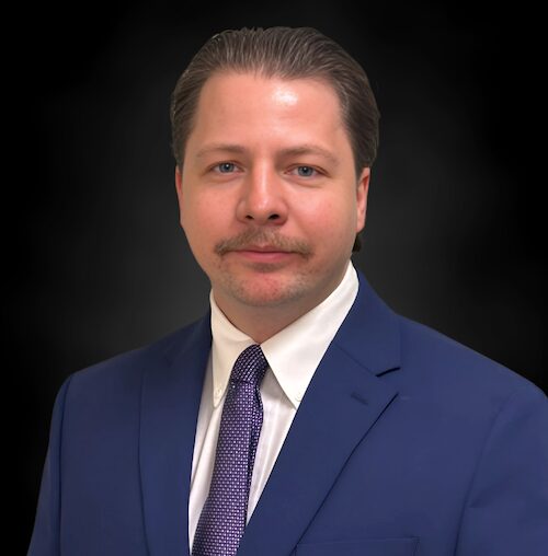A man with light skin and slicked-back brown hair is wearing a blue suit, white shirt, and purple tie, standing against a plain dark background. He has a mustache and a neutral expression.