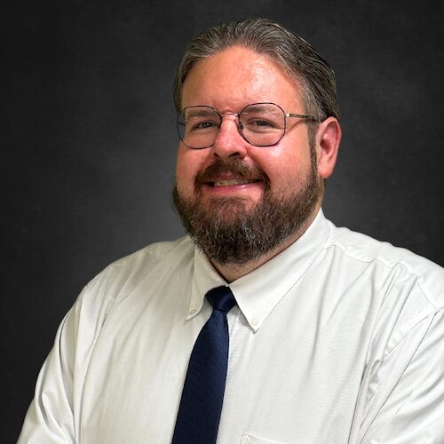 A smiling man with glasses, a beard, and mustache, wearing a white dress shirt and a dark tie, poses against a plain dark background.