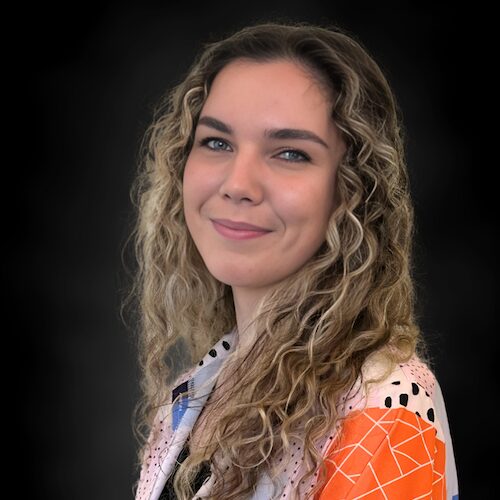A young woman with long, curly blonde hair smiles slightly, wearing a patterned blouse with orange, white, and black colors, set against a dark background.