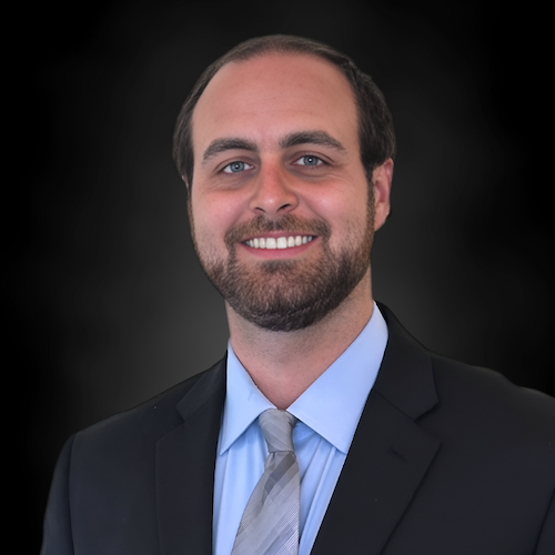 A man with light skin, short dark hair, and a beard is smiling, wearing a black suit jacket, light blue shirt, and light gray tie, posed in front of a dark, gradient background.