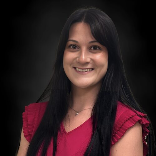 A woman with long black hair, wearing a sleeveless red top with ruffled edges, smiles at the camera against a dark, blurred background.