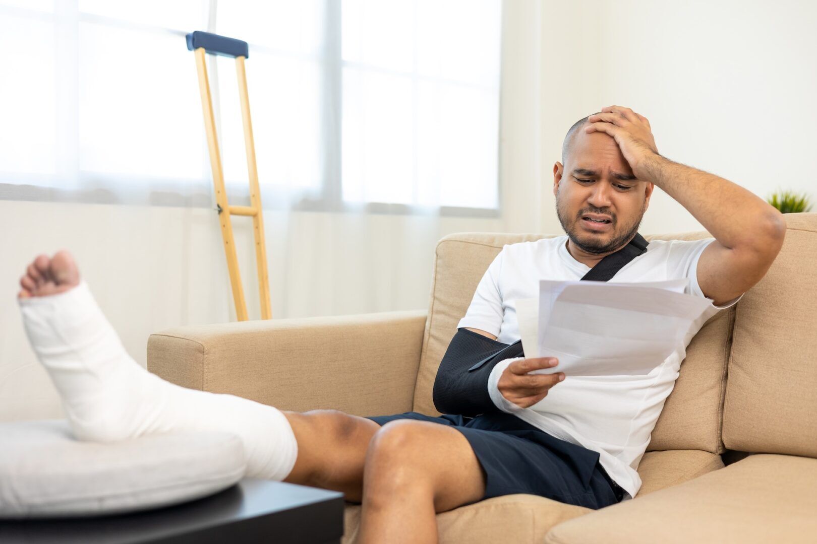 A man with a cast on his leg and his arm in a sling sits on a couch, looking stressed while reading a Guide To Managing Medical Expenses After a Car Accident. Crutches rest against the wall in the background.