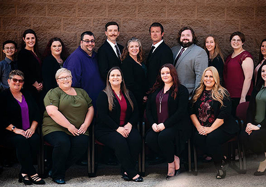 A group of fifteen adults, dressed in business casual attire, pose together in two rows in front of a brick wall. Some are seated while others stand behind, all smiling at the camera. A group of fifteen adults, dressed in business casual attire, pose together in two rows in front of a brick wall. Some are seated while others stand behind, all smiling at the camera.