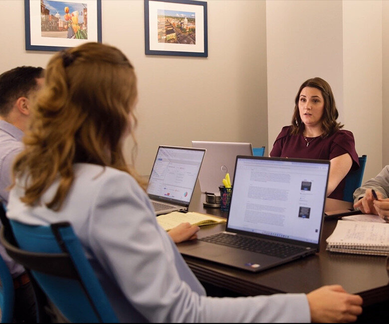 Four people are seated around a table in an office, engaged in a meeting. Laptops, notebooks, and office supplies are on the table. A woman in a maroon top appears to be speaking. Framed photos hang on the wall behind her.