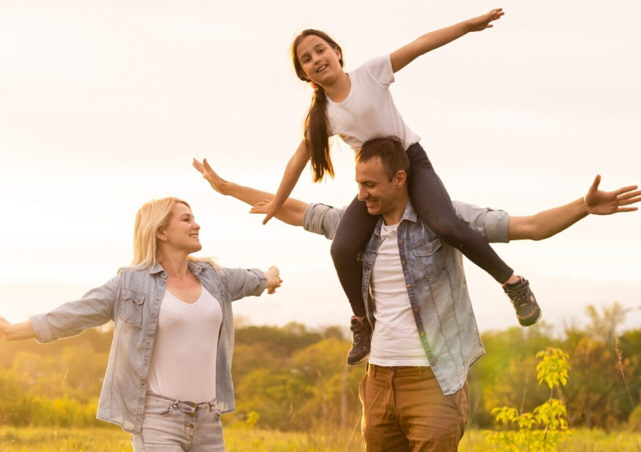 A smiling family of three stands outdoors in a field at sunset. The father carries a young girl on his shoulders, both with arms outstretched. The mother stands beside them with arms open, all appearing joyful and playful.