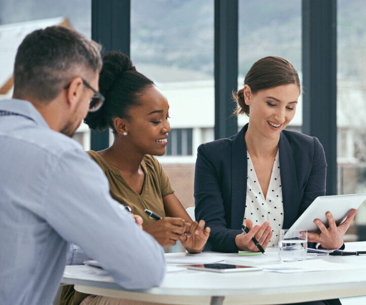 Three people sit at a table in an office, collaborating and smiling while looking at a digital tablet. Papers, pens, and a glass of water are on the table, and large windows show an outdoor view.