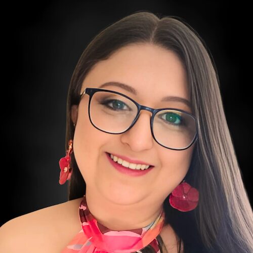 A woman with long brown hair and glasses smiles at the camera. She is wearing red floral earrings and a pink patterned top, set against a plain black background.