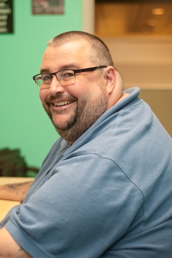 A man with short hair, glasses, and a beard smiles while sitting at a desk. He is wearing a blue polo shirt and is pictured in an indoor setting with mint green walls.