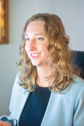 A woman with curly, light brown hair and a light blue blazer smiles as she looks at something off camera. She is seated indoors in a softly lit room.