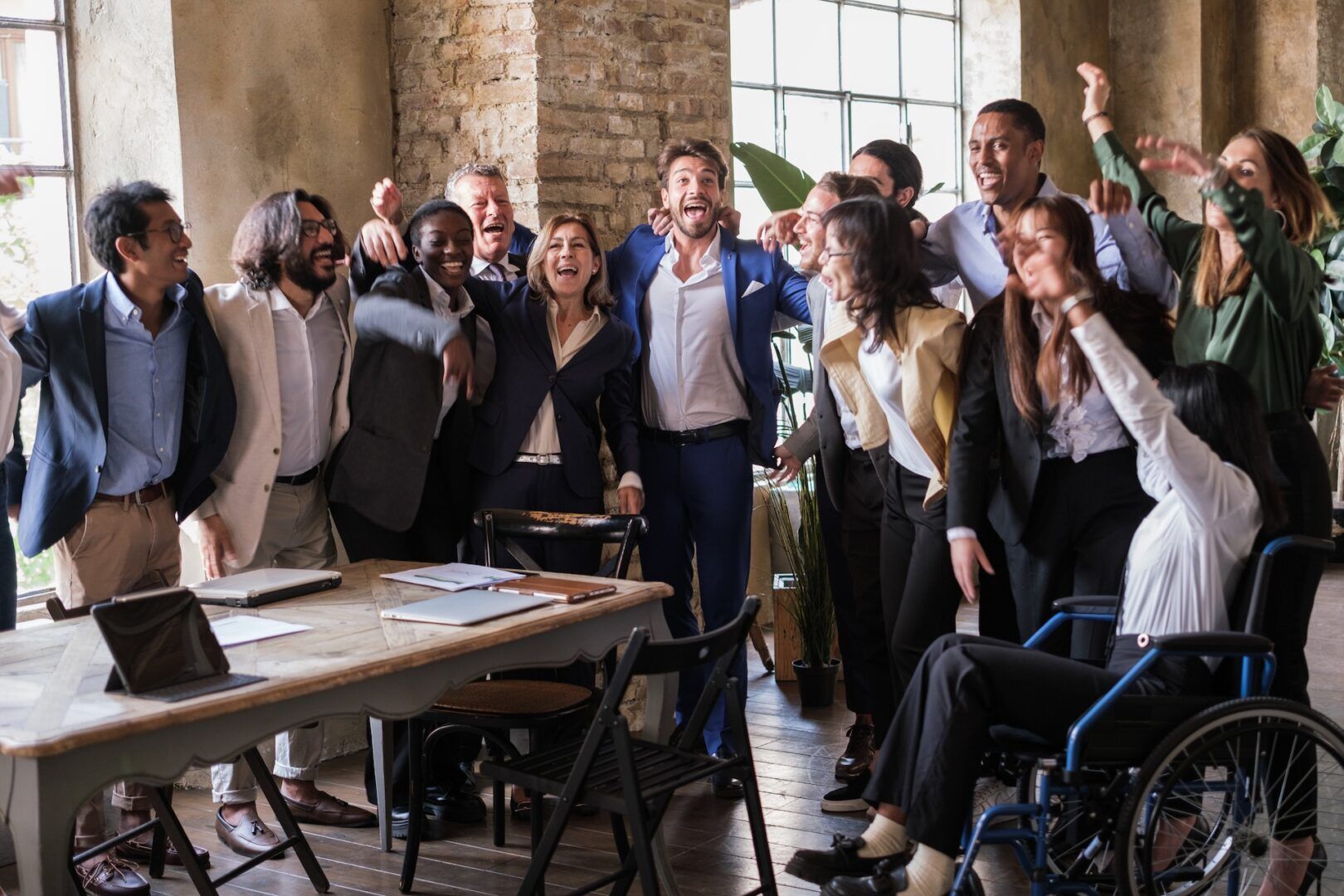 A diverse group of professionals, including one person in a wheelchair, stand together in an office, smiling and celebrating New Workplace Rights for WV Employees in 2025 around a table with notebooks and papers. Sunlight streams through large windows behind them.