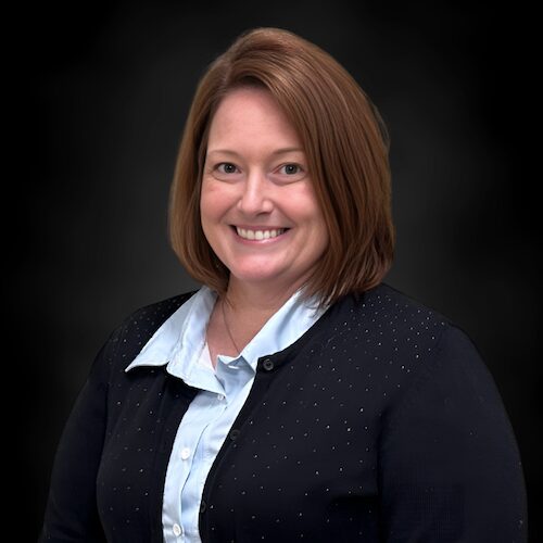 A woman with straight, shoulder-length brown hair smiles at the camera. She is wearing a light blue collared shirt under a dark, textured sweater. The background is plain and dark.