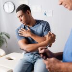 A man sits on an exam table holding his shoulder in pain while a healthcare professional in scrubs writes on a clipboard, perhaps discussing concerns like "Did Nepotism Lead To Your Wrongful Termination? Your Rights in WV & Ohio Explained.