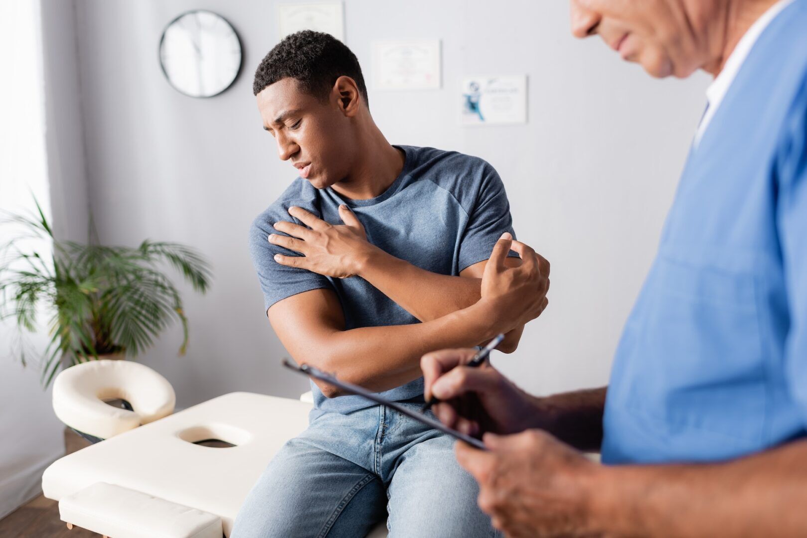 A man sits on an exam table holding his shoulder in pain while a healthcare professional in scrubs writes on a clipboard, perhaps discussing concerns like "Did Nepotism Lead To Your Wrongful Termination? Your Rights in WV & Ohio Explained.