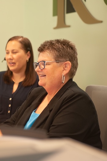 Two women sitting indoors at a desk, one in focus smiling and wearing glasses and a black blazer, the other slightly blurred and wearing a headset. A large letter “R” is visible on the wall behind them.
