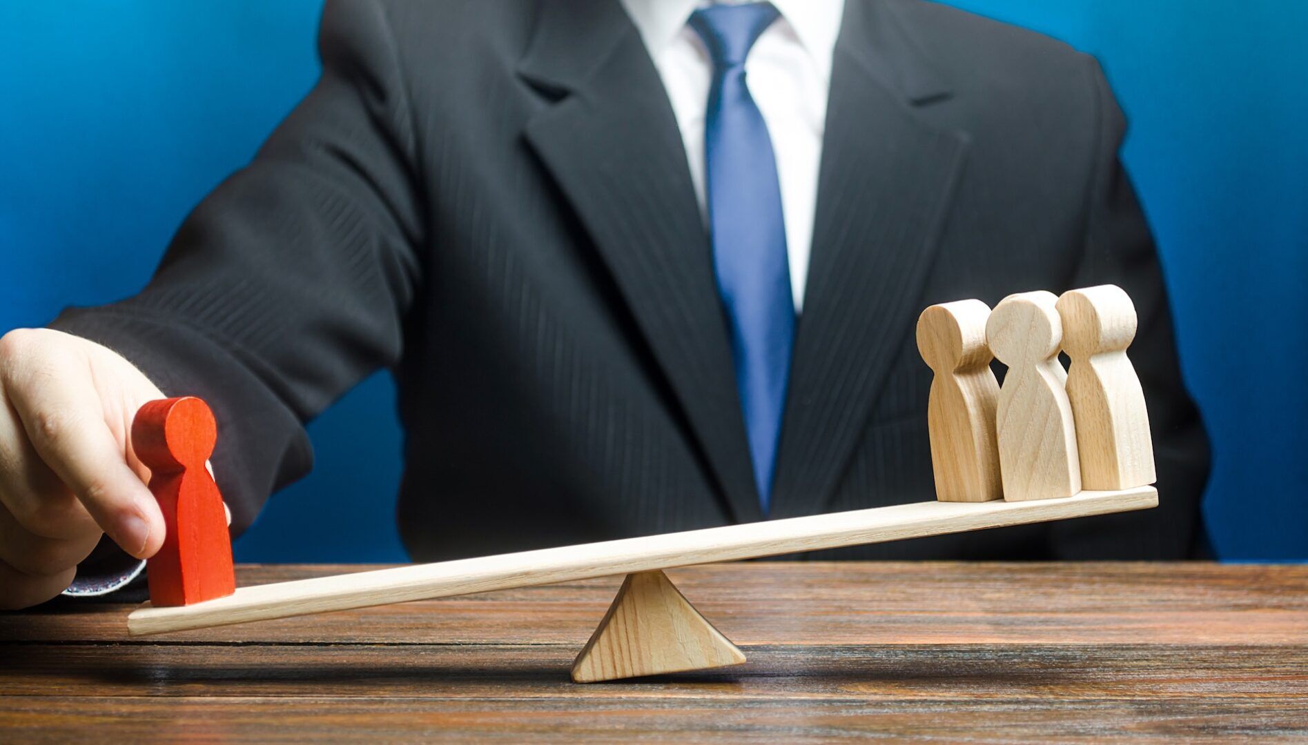 A person in a suit balances a wooden seesaw with one red figure on one side and three natural wood figures on the other side, symbolizing decision-making or influence of the individual versus a group.