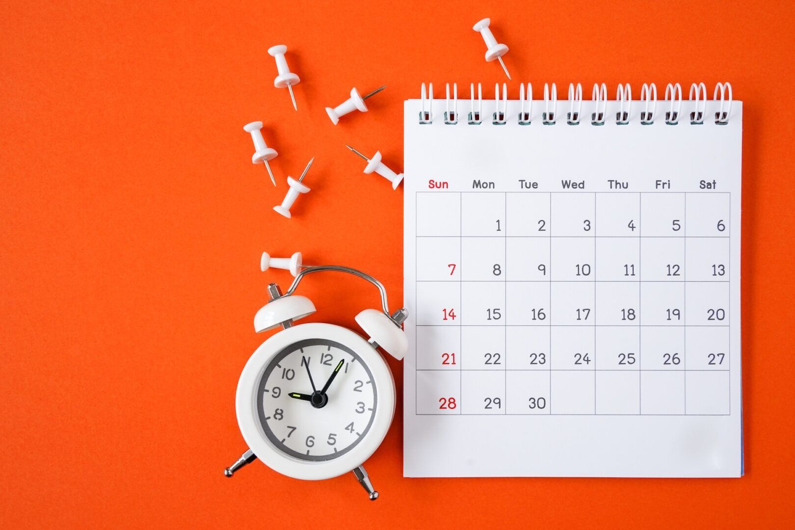 A white alarm clock, a spiral calendar open to a month view, and several white push pins are arranged on an orange background—reminding you to learn if nepotism led to your wrongful termination in WV & Ohio and what your rights are. The clock shows 10:12.