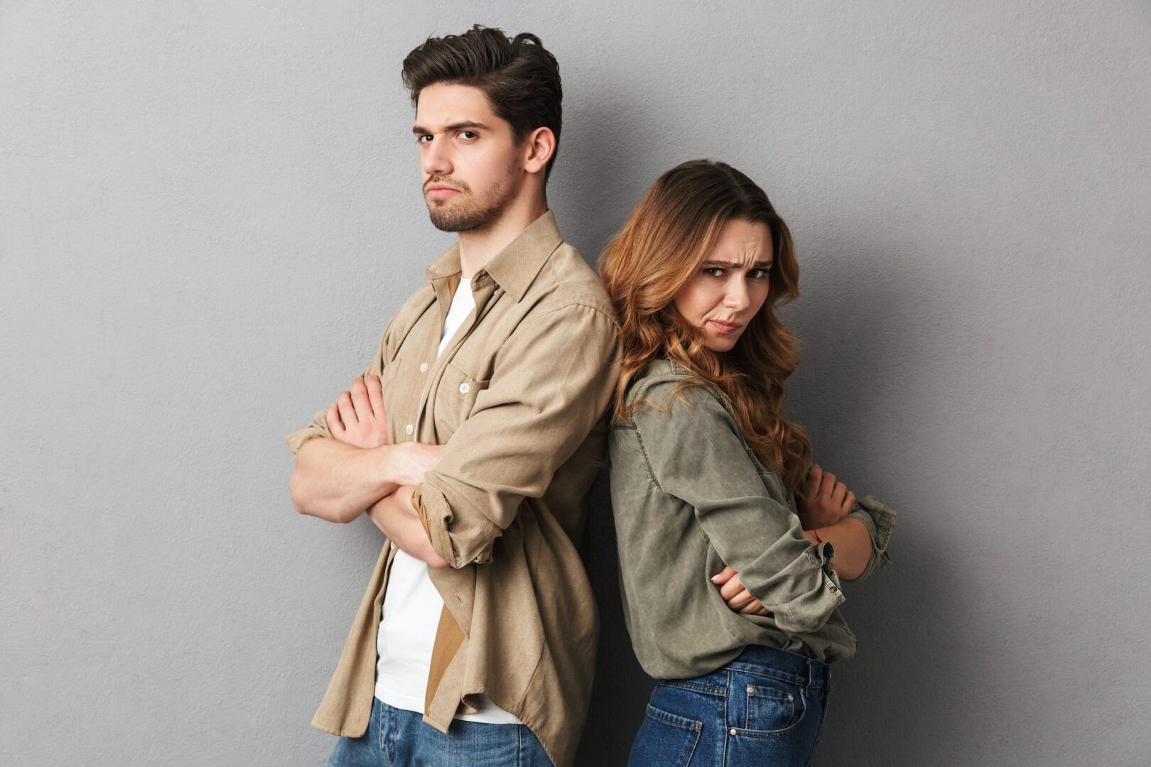 A man and woman stand back-to-back with arms crossed, both looking upset and serious against a plain gray background—capturing the tension that may signal Is Your Relationship in Trouble? Common Divorce Signs in West Virginia & Ohio.