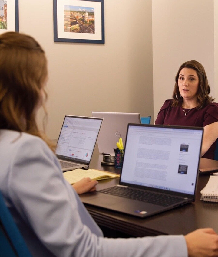 Two women sit across from each other at a table in an office, both with open laptops and notebooks. One woman faces the camera while the other is seen from behind. Framed pictures hang on the wall.