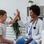 A smiling doctor in a white coat gives a high-five to a young boy in a medical office, where child costs—like medical expenses—can raise questions such as, Who Pays for What After Divorce? Both appear happy and relaxed as the doctor holds a small device.