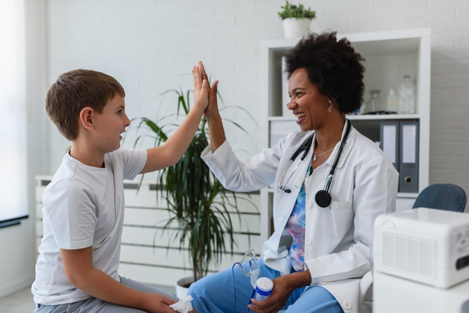 A smiling doctor in a white coat gives a high-five to a young boy in a medical office, where child costs—like medical expenses—can raise questions such as, Who Pays for What After Divorce? Both appear happy and relaxed as the doctor holds a small device.