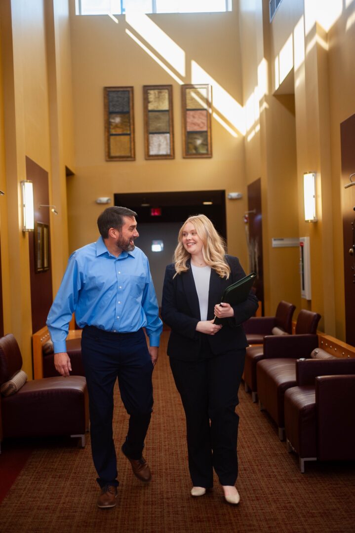 A man in a blue shirt and a woman in a black suit walk and smile at each other in a brightly lit hallway with yellow walls and brown chairs. A man in a blue shirt and a woman in a black suit walk and smile at each other in a brightly lit hallway with yellow walls and brown chairs.