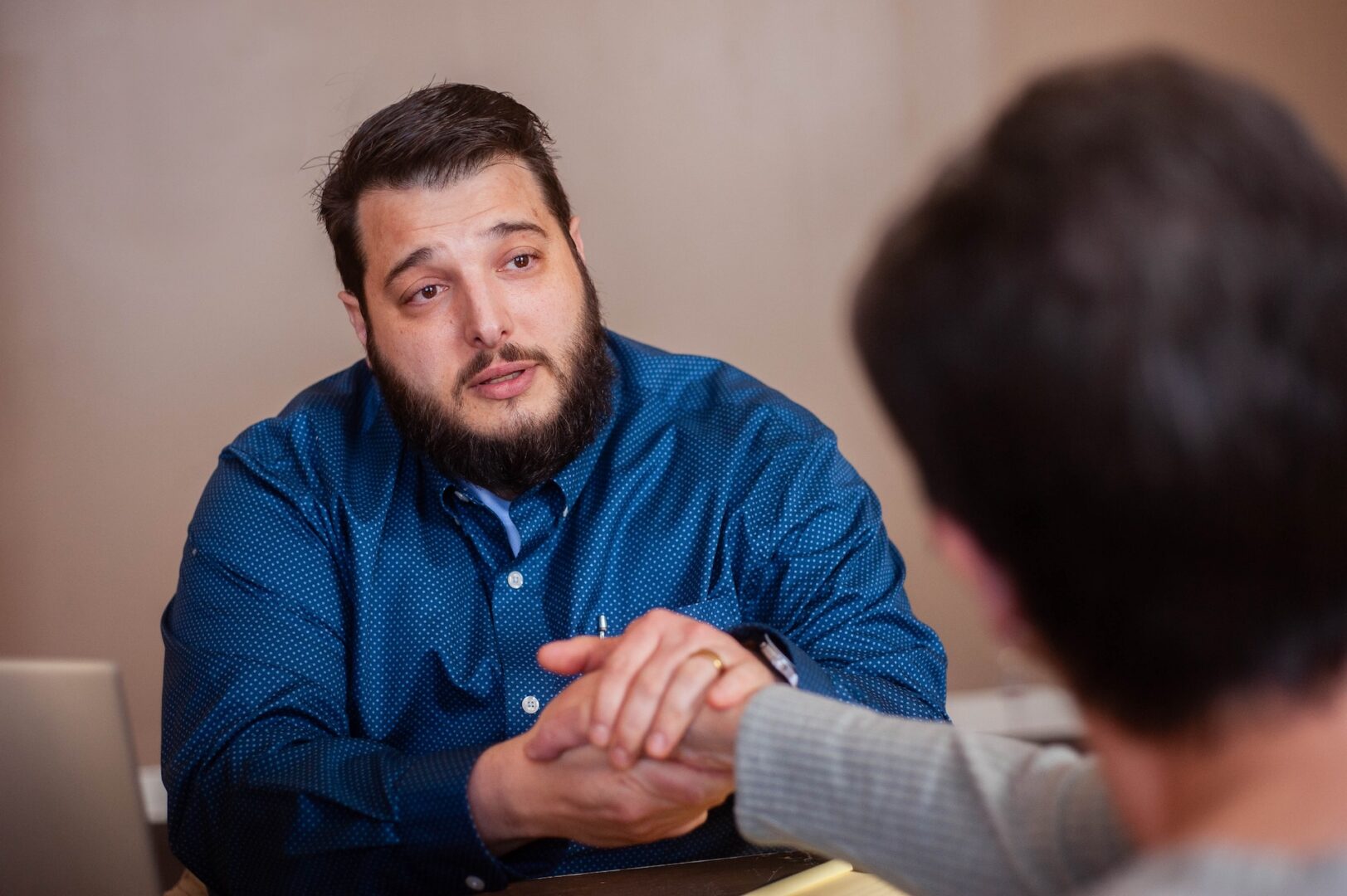 A man with a beard in a blue shirt sits at a desk, holding hands with another person across from him in a comforting gesture, showing support and empathy in a conversation. A man with a beard in a blue shirt sits at a desk, holding hands with another person across from him in a comforting gesture, showing support and empathy in a conversation.