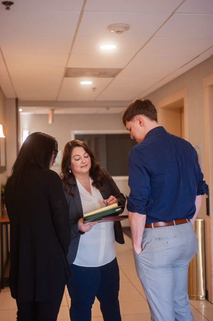 Three people stand in a hallway having a discussion. One woman holds an open folder and talks to the other two, who are listening attentively. The environment appears to be professional and well-lit. Three people stand in a hallway having a discussion. One woman holds an open folder and talks to the other two, who are listening attentively. The environment appears to be professional and well-lit.