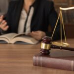 A gavel rests on a law book beside scales of justice, as two people in business attire discuss how to use West Virginia's two-year personal injury statute of limitations while reviewing an open book in the background.