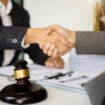 Two people in business suits shake hands across a desk with a judge’s gavel, a contract, and documents, suggesting that getting divorced for the first time? Discover the benefits of going uncontested for a smooth legal agreement.