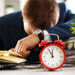 A person in a suit sits at a desk, looking tired, surrounded by an open book, a laptop, papers, and an alarm clock—perhaps overwhelmed by questions like "Who Pays for What After Divorce? Child Costs Explained." A green plant adds a touch of calm.