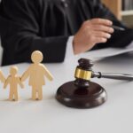 A judge in a black robe sits at a desk holding papers, with wooden family figures and a gavel in the foreground, symbolizing how courts decide custody and what factors WV courts consider in child custody cases.