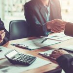 Three people in business attire sit at a desk with documents, a calculator, and pens. Two shake hands as the third claps, highlighting the importance of addressing Workplace Sexual Harassment: Signs, Evidence & Legal Steps for safe collaboration.