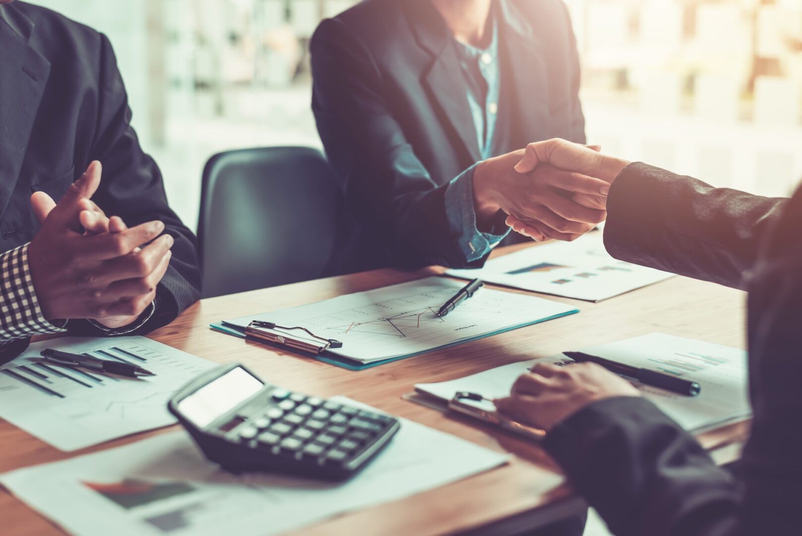 Three people in business attire sit at a desk with documents, a calculator, and pens. Two shake hands as the third claps, highlighting the importance of addressing Workplace Sexual Harassment: Signs, Evidence & Legal Steps for safe collaboration.